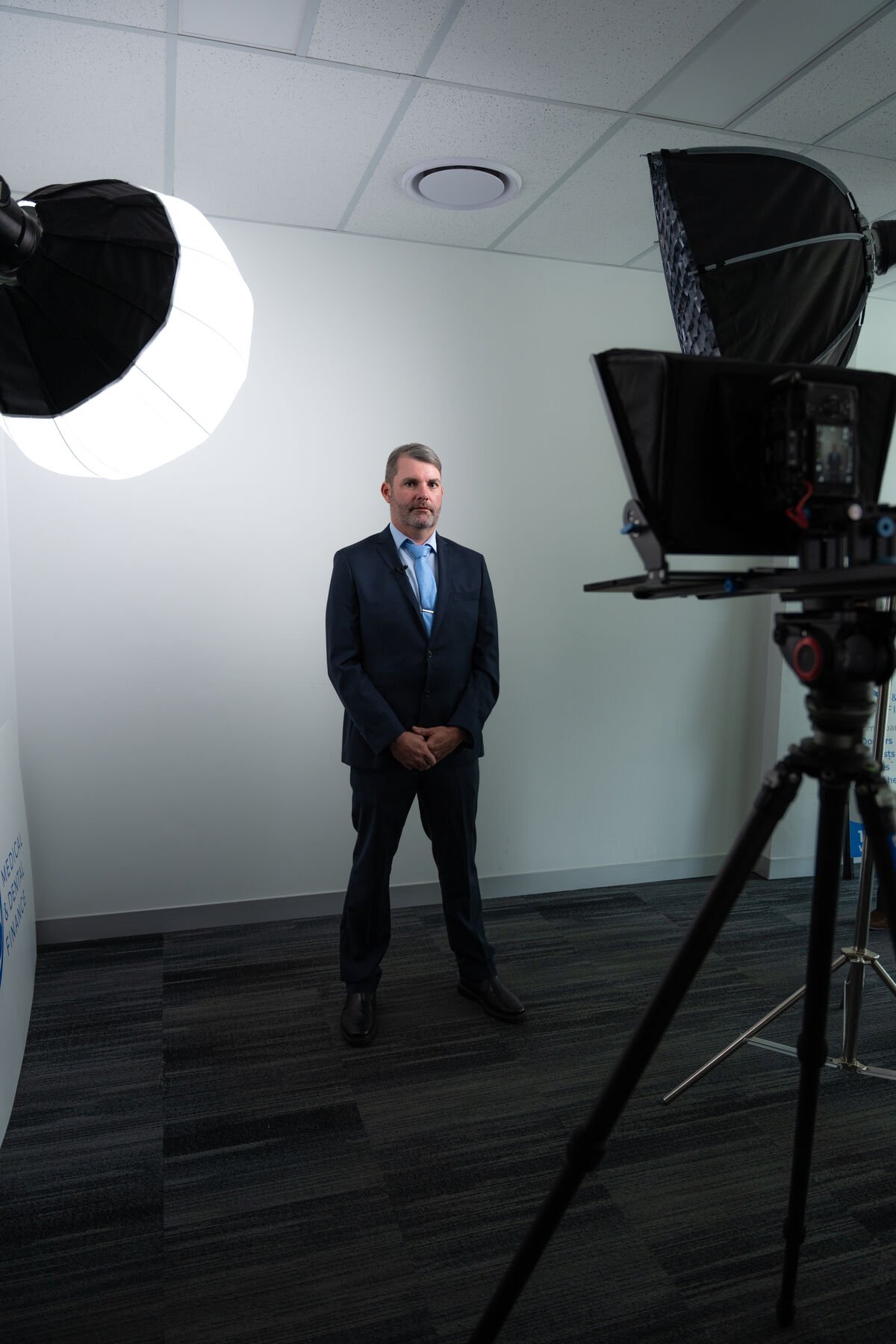 Studio portrait of a suited subject lit with a softbox key, teleprompter rig visible on the right
